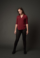 full length portrait of brunette girl wearing  red shirt and leather pants. standing pose, on grey studio background.