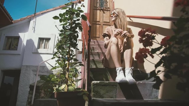 Beautiful and happy female waiting for a friend sitting in stairs of adorable small Mediterranean house street of small town of Europe feeling happy and enjoying sun in a summer vacations and holidays