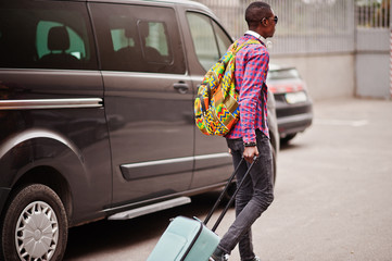 African american man in checkered shirt, sunglasses and earphones with suitcase and backpack. Black man traveler against van car.