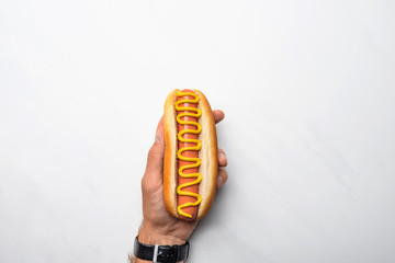 cropped shot of man holding tasty hot dog poured with mustard on white marble surface