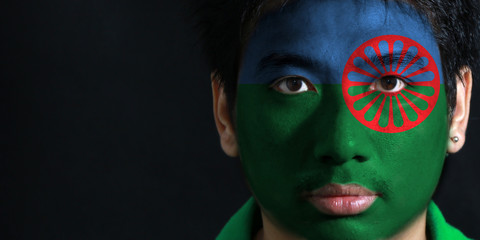 Portrait of a man with the flag of the Romani people painted on his face on black background. blue...
