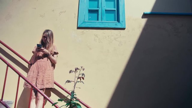 Beautiful and happy feemale in a nice dress is waiting and texting by smartphone app under window street of small port town of Europe enjoyng the sun good wether in a summer in vacation and holidays