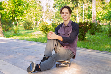 Obraz premium Young man sitting and relaxing on skateboard after skating in city park. Teen boy skater
