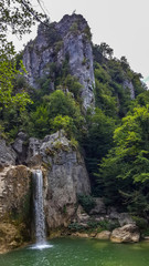 A green pond formed by a waterfall flowing through the rocks reaching into the sky