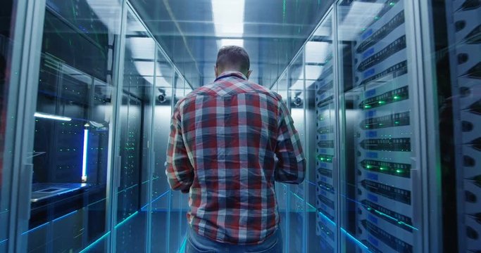 Back view of casual man with tablet walking among server racks in data center room looking at hardware