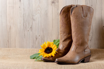 A pair of woman's brown cowboy boots with a bright yellow sunflower on acanvass with wooden...