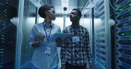 Coworking African American man and woman with tablet walking in corridor with server racks in data center checking hardware in team - Powered by Adobe