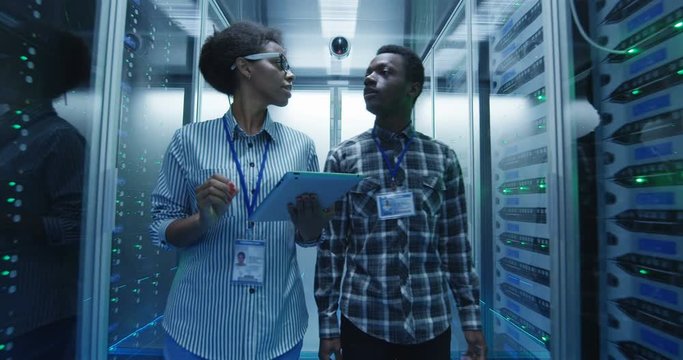 Coworking African American man and woman with tablet walking in corridor with server racks in data center checking hardware in team