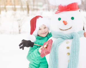 portrait of smiling boy in red christmas hat holds gift box and hugs snowman