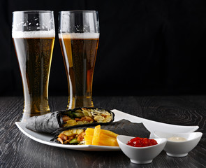 Beer in a glass and shawarma in pita bread is cut and lies on a wooden surface. The Middle Eastern dish is prepared on the grill and served with sauce.
