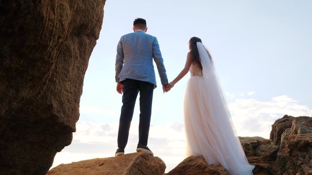 Unrecognizable wedding couple holding hands on sunset or sunrise background. Bride and groom standing outdoor near sea. Sun flares. Slow motion