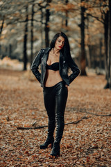 Beautiful dark haired young woman wearing a black bra, leather jacket and black leather pants posing in the woods on a sunny autumn afternoon.