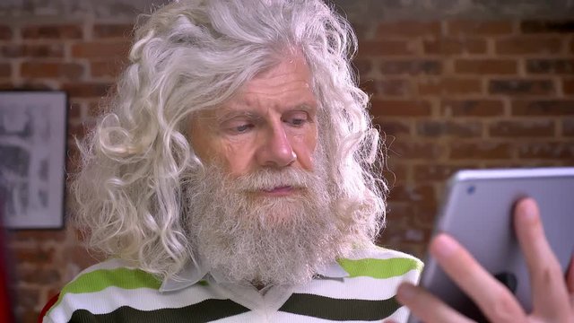 Ancient caucasian male with awesome white beard and lang bulky hair is typing on his tablet in modern brick office, casual lifestyle