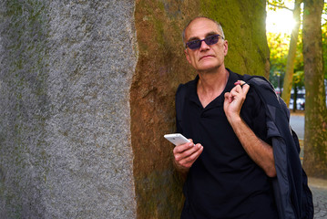 attraktiver europ&auml;ischer Mann mittleren Alters mit kurzen Haaren, Sonnenbrille, schwarzem T-Shirt  lehnt mit nachdenklichem Blick an einem Felsblock und hat ein Smartphone in der Hand