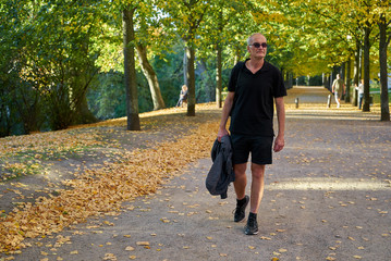Fototapeta premium attraktiver europäischer Mann mittleren Alters mit kurzen Haaren, Sonnenbrille, schwarzem T-Shirt und Shorts spaziert durch einen herbstlichen Park