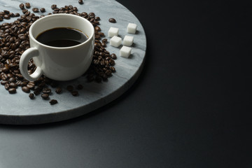 a cup of coffee with a pile of coffee beans and the sugar cubes on the round marble tray at the black table.
