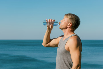 A man drinking and pours water on his face from bottle on the ocean, refreshing after a workout