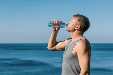 A man drinking and pours water on his face from bottle on the ocean, refreshing after a workout