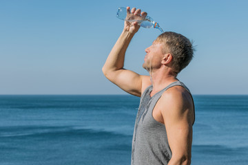 A man drinking and pours water on his face from bottle on the ocean, refreshing after a workout