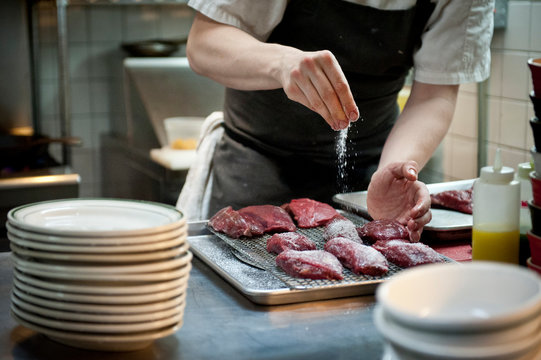 Chef Sprinkling Salt On Meat In A Restaurant Kitchen