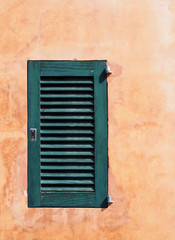 Fototapeta na wymiar a small green painted closed wooden shutter on an ochre orange old rough textured wall in bright sunlight and shadow