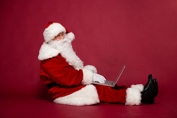 Happy Santa Clause working on laptop while sitting on red background, Christmas and New year concept