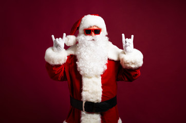 Stylish man in Santa Clause costume and sunglasses looking at camera and showing rock sign gesture...