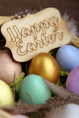 Wicker basket with colored Easter eggs, feathers, decorative chicken and a wish on a wooden label on a dark wooden background. Focus on the golden egg.