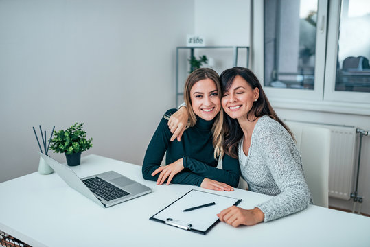 Two Casual Business Women Hugging While Sitting At Work. Friends Working Together.