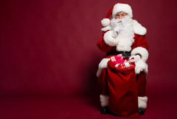 Man in Santa Clause costume sitting with gift sack on red background, Christmas and New year concept
