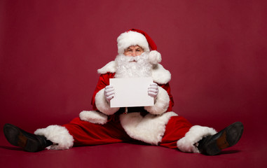 Funny man in Santa Clause costume with white blank card in hand sitting on red background, Christmas and New year concept