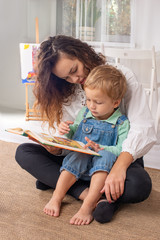 Young mother or nanny with small child boy sit on the floor on a rug in the room at home and read a...