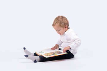 Full length portrait of a little serious boyl wearing  white shirt and blask pants.  He is sitting on the floor in profile and is considering a children's book on a white background in the studio. © rozaivn58