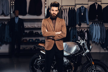 Obraz na płótnie Canvas Elegantly dressed man with stylish beard and hair posing near retro sports motorbike at the men's clothing store.
