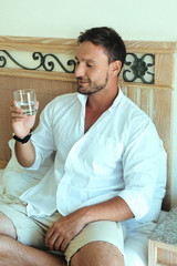 happy young handsome bearded man in white clothes enjoying his morning with cup of coffee or water smiling. Bachelor holding a glass of water, sitting on a bed, he is a freelancer