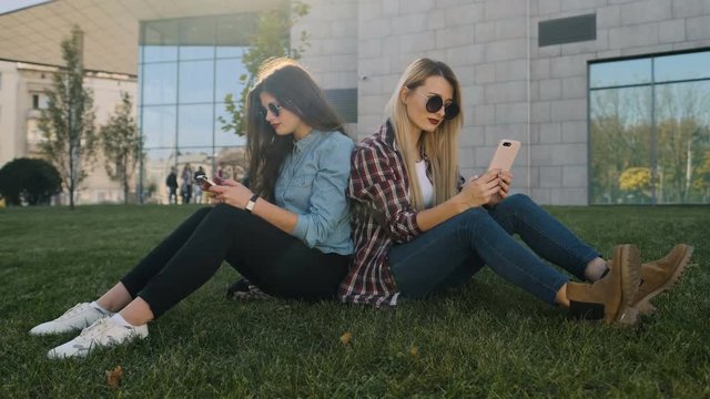 Two attractive hipster girls in casual clothing relax and chat over. Woman using smart phone typing text message outside. They are happy and smiling, sitting on the grass
