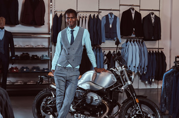 Fototapeta na wymiar Elegantly dressed African American young man posing near retro sports motorbike at the men's clothing store.