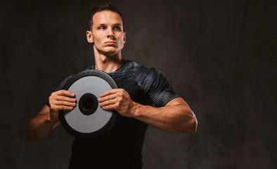 A young man in sportswear holding a barbell disk on dark background.