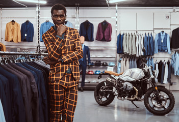 Fototapeta na wymiar Elegantly dressed African man posing with hand on chin while standing in a classic menswear store.