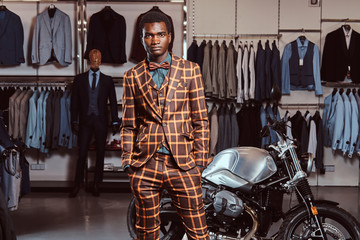 Fototapeta na wymiar African-American man dressed in a trendy elegant suit posing with hands in pockets near retro sports motorbike at the men's clothing store.