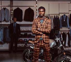 African-American man dressed in a trendy elegant suit posing with crossed arms near retro sports motorbike at the men's clothing store.