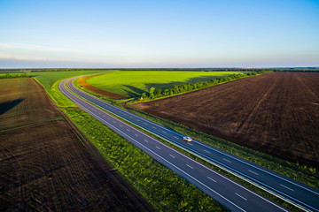 White car driving on asphalt road along the green fields in rural landscape. Road seen from the air. Aerial view landscape. drone photography.