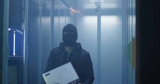 Anonymous man in mask using laptop in smoky spark data center room hacking and destroying servers while starting fire