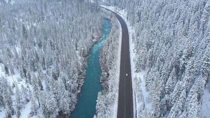 AERIAL: White car drives down empty road running along the river in the winter.