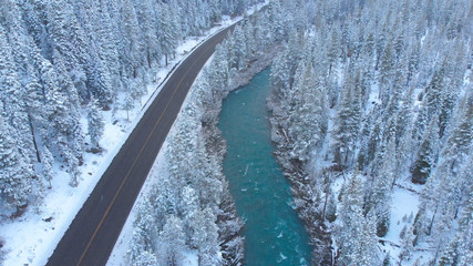 AERIAL: Flying over emerald river and scenic road running through snowy forest.