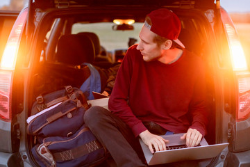young freelancer website creator sitting in the car trunk with laptop and working in the trip f