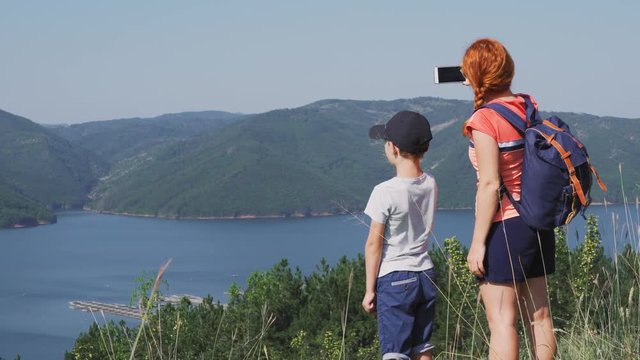 Panorama of beautiful mountains. Tourists, mother and son, with backpacks make a walk through the mountainous terrain and film a green horizon of mountains, a lake on a smartphone. Family on a tourist