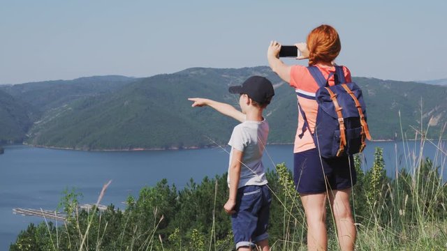 Panorama of beautiful mountains. Tourists, mother and son, with backpacks make a walk through the mountainous terrain and film a green horizon of mountains, a lake on a smartphone. Family on a tourist