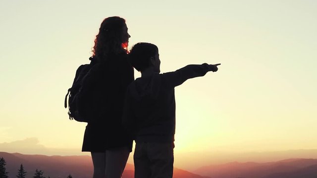 A slide of a woman and her son stand on top of a mountain against the background of a mountain range, look at the horizon, raising their hands up. Silhouettes of tourists at sunset in the orange light