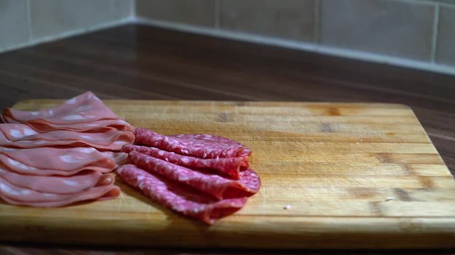 A man prepares a meat and cheese platter by placing ham, salami, and cheese on a wooden serving tray, filmed in UHD 4K